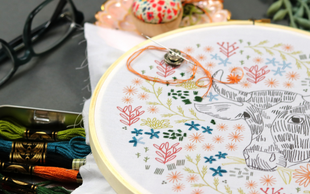 embroidery hoop art with cow and flowers displayed on a desk with embroidery floss, a pin cushion and eyeglasses.