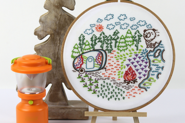 embroidered hoopart with fun camping scene displayed on an easel with camp lantern and wooden tree bookend  