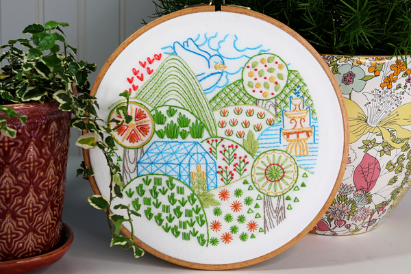 embroidery hoop art with summer landscape scene displayed alongside house plants in ceramic pots