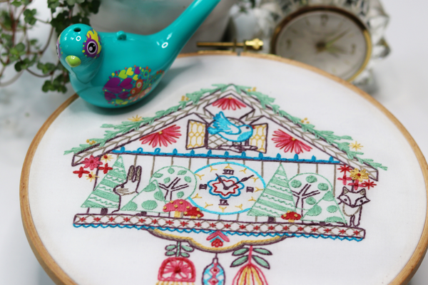 embroidered hoop art with vintage cuckoo clock displayed with bird figurine, old clock and house plant.