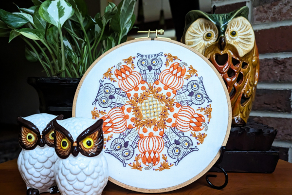 Embroidered hoop art of pumpkins and owls displayed on stand.