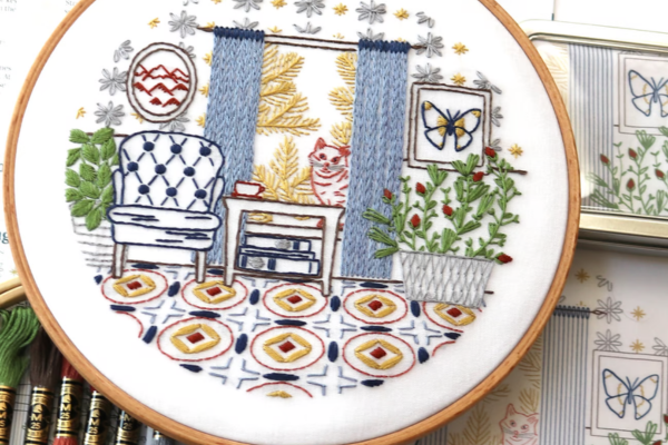 Finished embroidery hoop-art of sitting room with chair, drapes, rug and plants.
